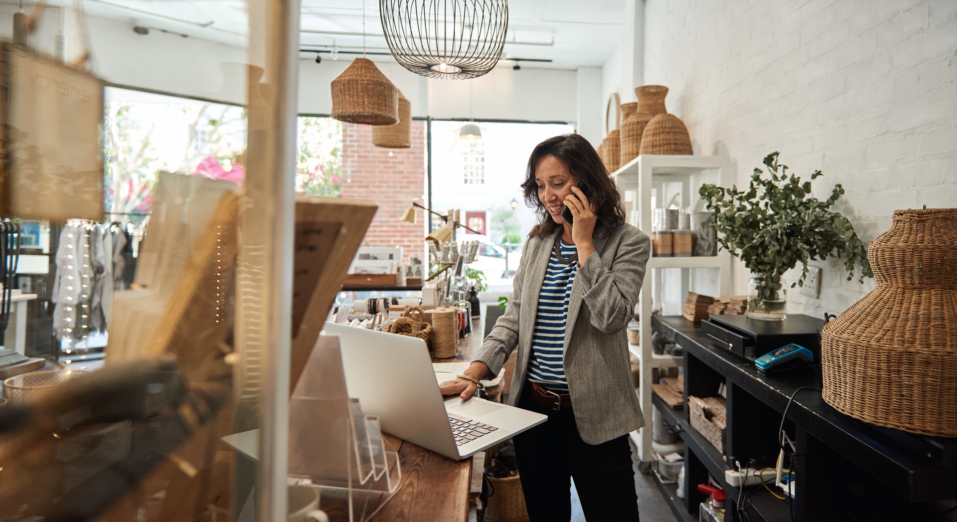 Smiling woman standing behind a counter in her stylish boutique working on a laptop and talking on a phone